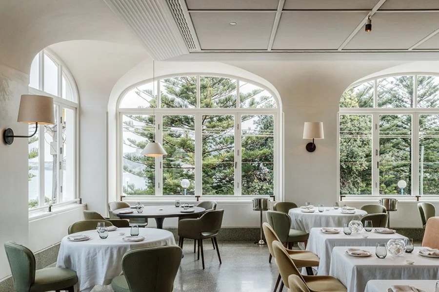 A bright, airy restaurant with large arched windows showing trees outside, white walls, and round tables with white tablecloths and beige and green chairs.