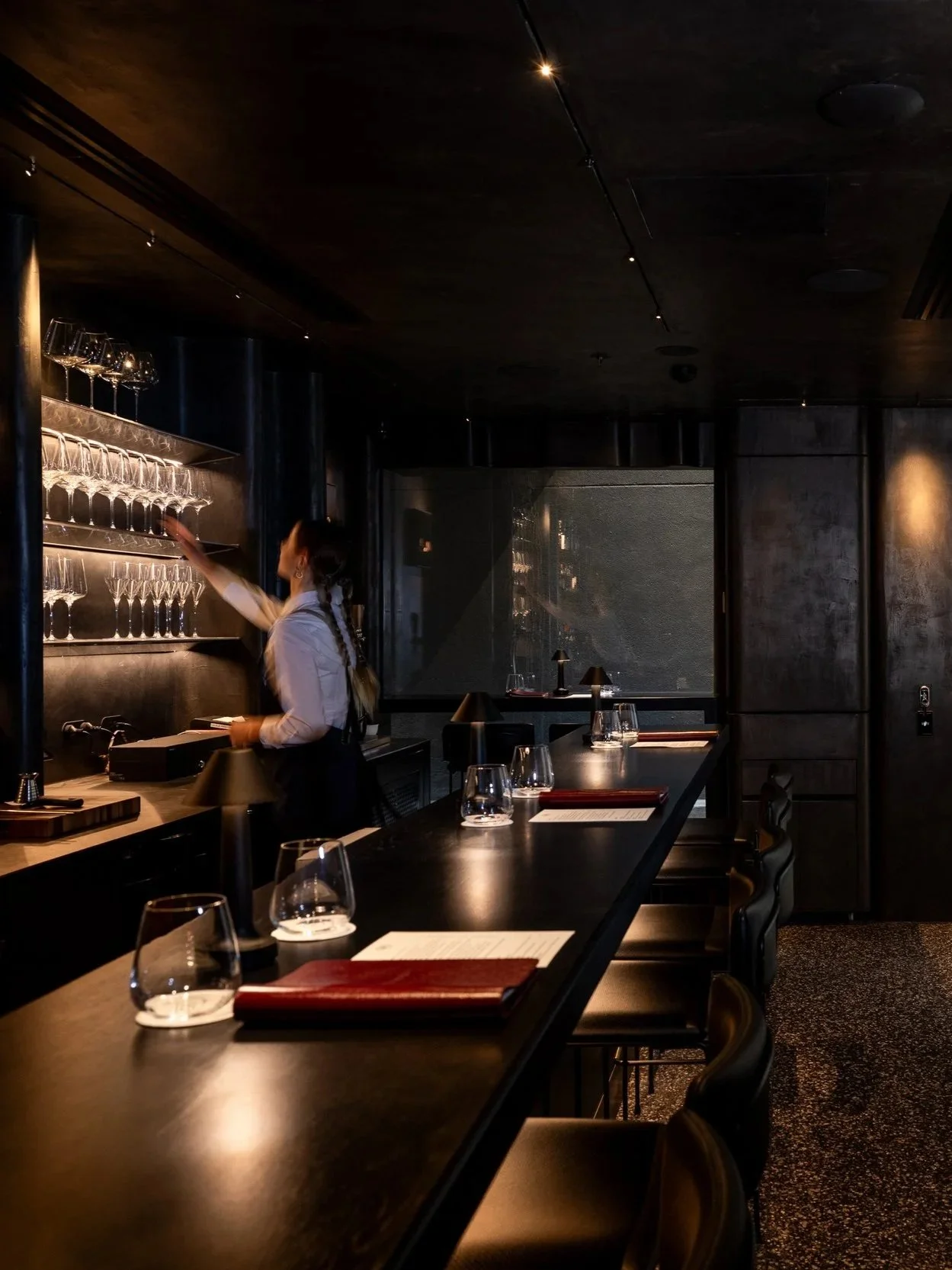A dimly lit restaurant interior with a bar counter and drinks, a bartender reaching for glasses on a shelf, and place settings with glassware and menus on the tables.