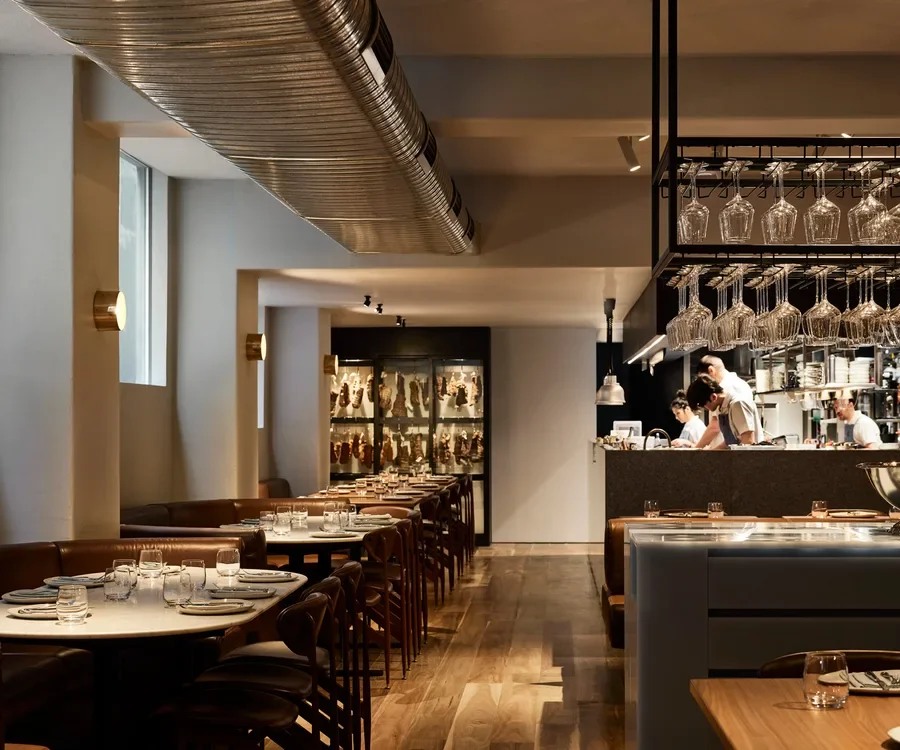 Inside a modern restaurant with neatly arranged tables, leather chairs, and a visible open kitchen area with staff preparing food.