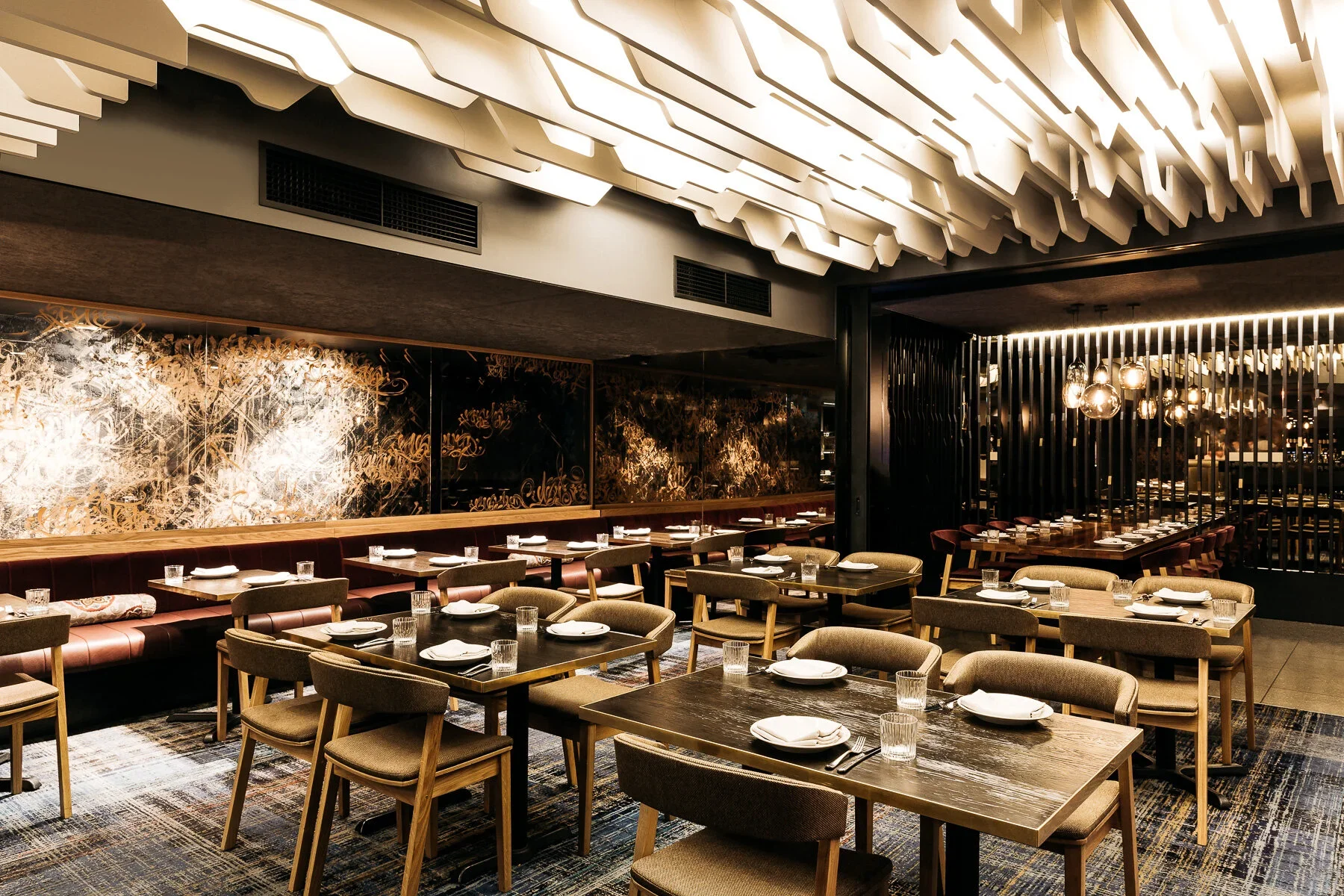 Elegant dining room with wooden tables set with white plates, glasses, and napkins, featuring a modern ceiling design and artistic wall decor in a restaurant.