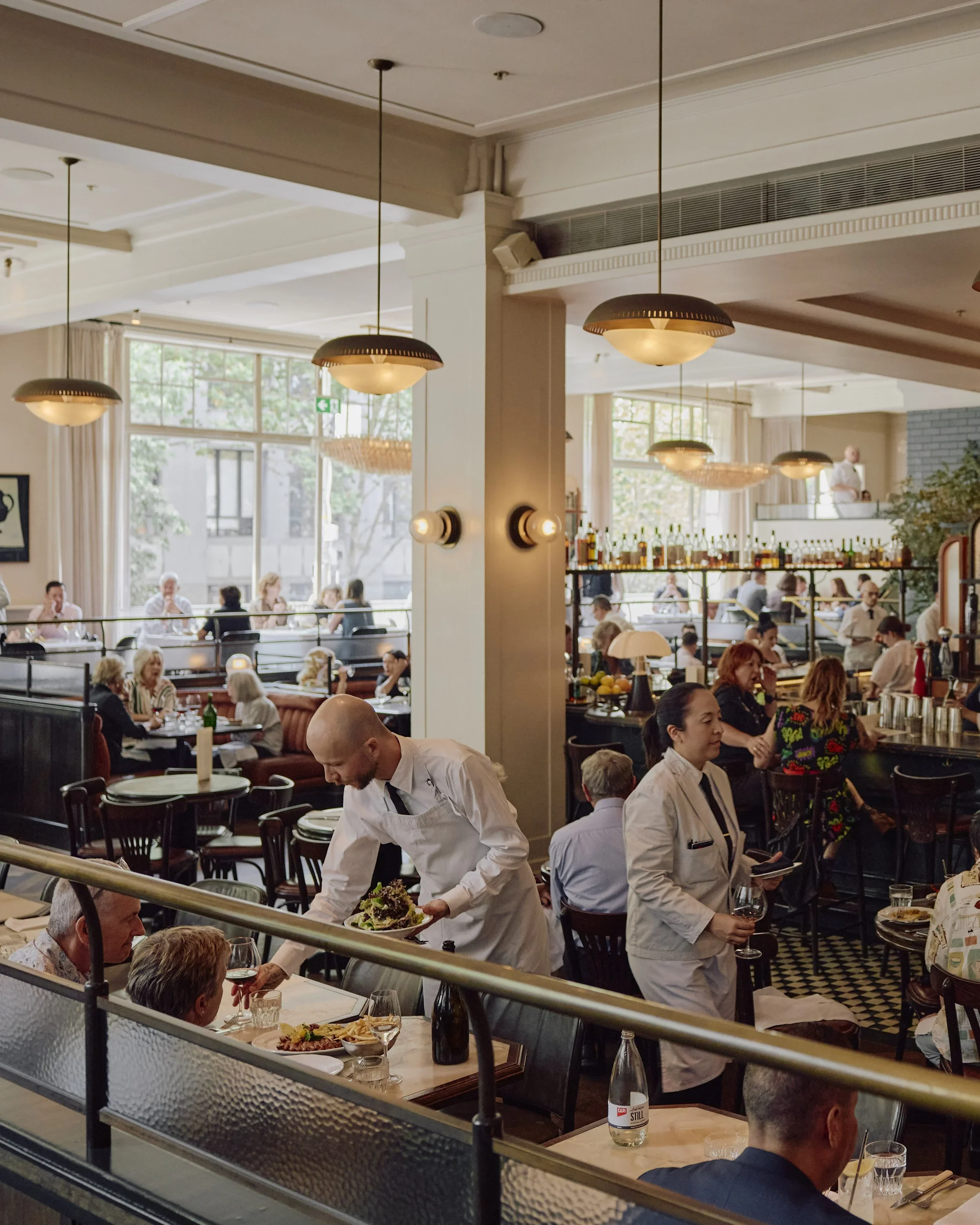 The image depicts a busy restaurant interior with several patrons dining and a waitress serving food. The decor features hanging pendant lights, large windows, and a bar area in the background. The restaurant has a casual yet elegant atmosphere.