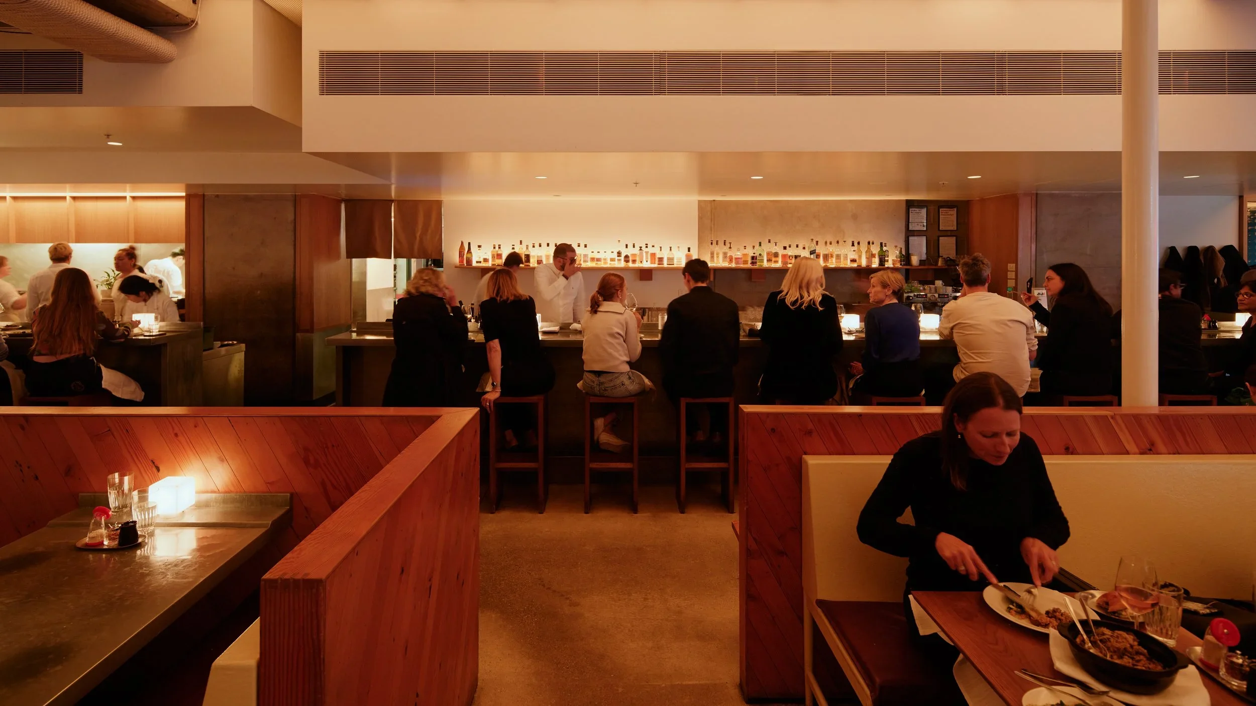 People sitting at a bar counter in a restaurant, with bottles of alcohol displayed on a shelf behind the bartender.