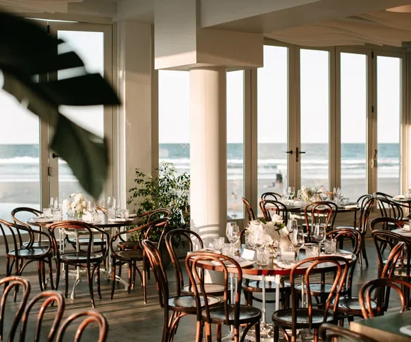 Beachside restaurant with round tables set with white floral centerpieces, glassware, and plates, featuring large windows overlooking the ocean.