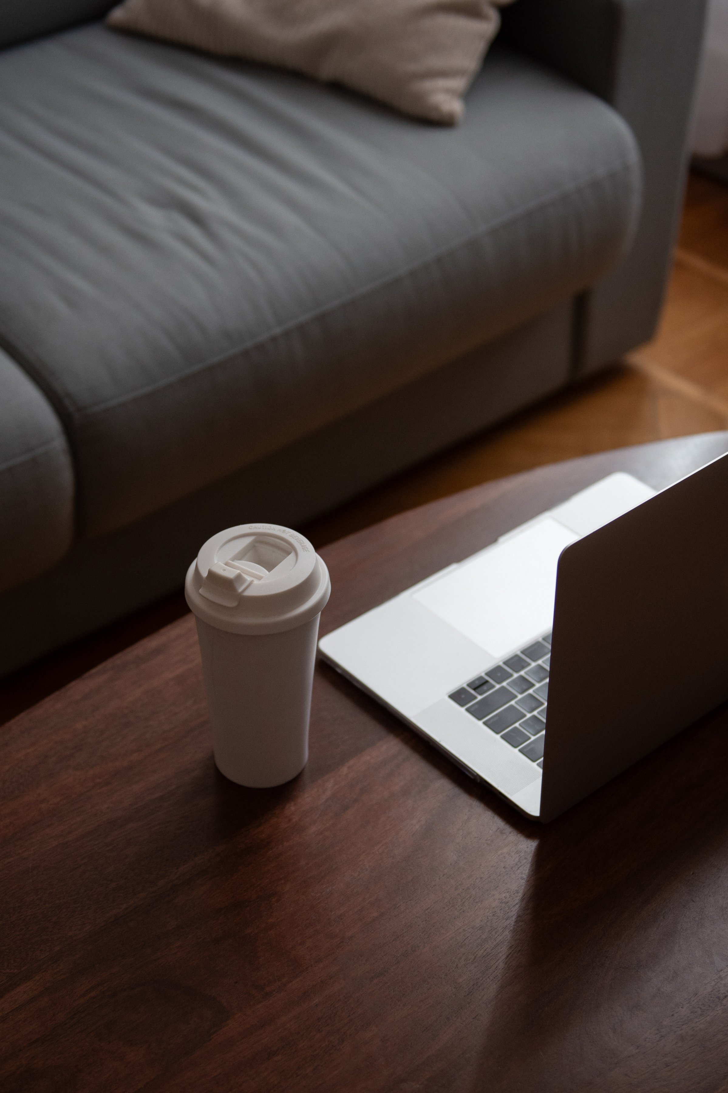 A desk with a white travel mug, a silver laptop, and a gray sofa with a beige pillow in the background.