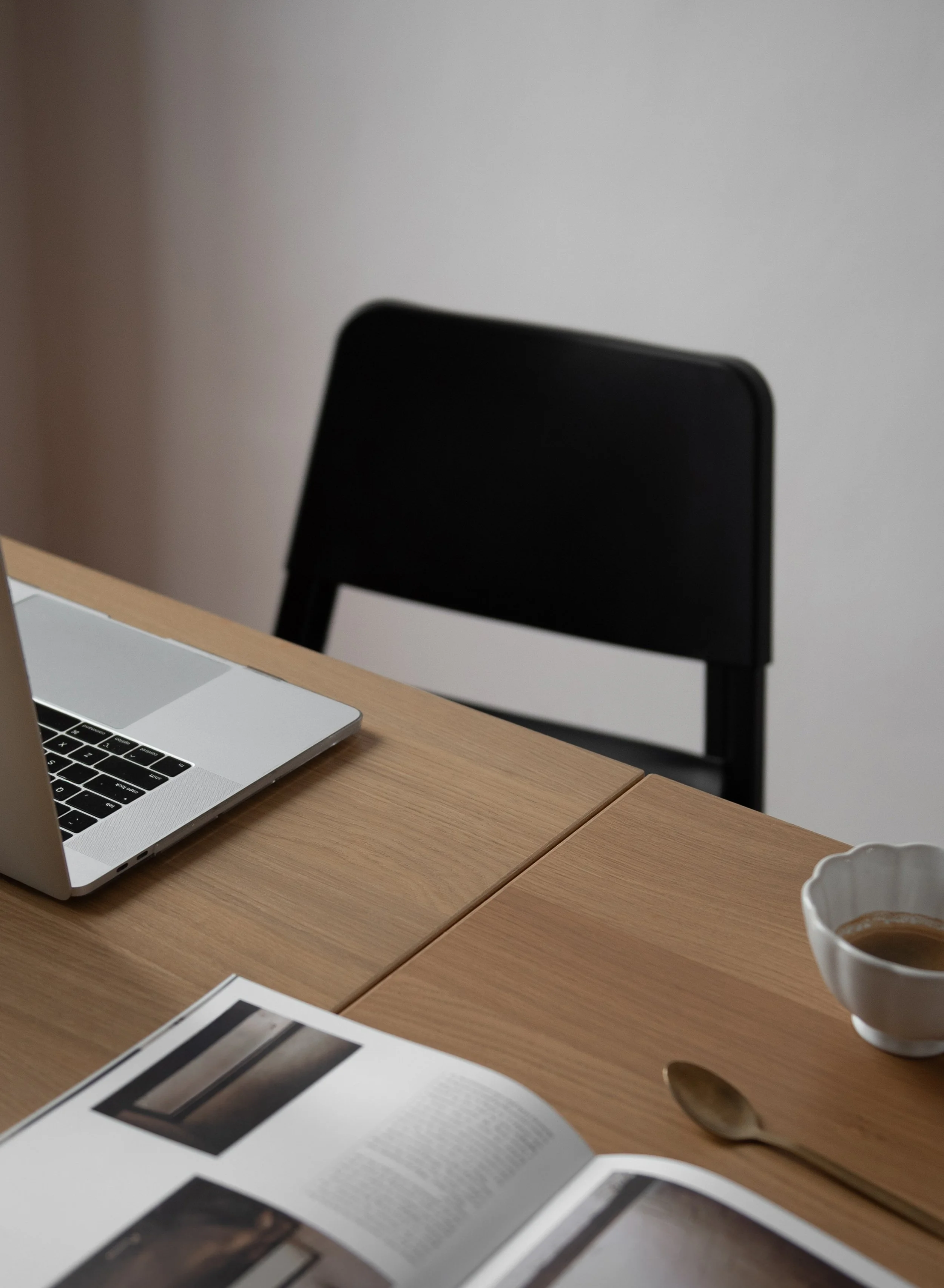 Part of a workspace with a wooden table, open magazine with images and text, a silver laptop, a spoon, a white cup with brown liquid, a blank white paper, and a black chair against a plain wall.