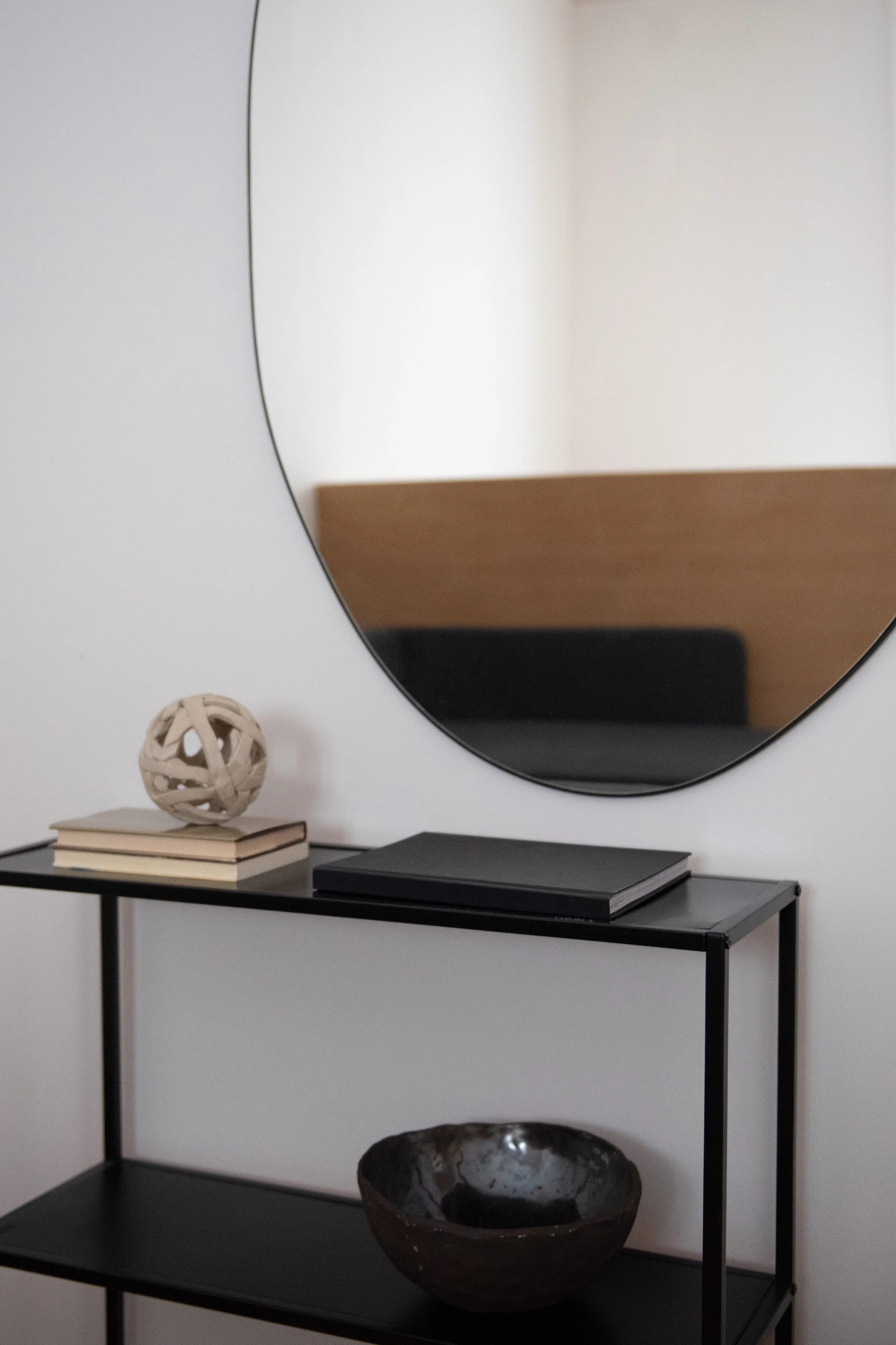 A black metal console table with three shelves against a white wall. On top of the table, there are three books, a decorative spherical sculpture, and a black rectangular object. The middle shelf holds a large dark ceramic bowl, and the bottom shelf appears empty. A round mirror hangs on the wall behind the table, reflecting part of a wooden piece of furniture.
