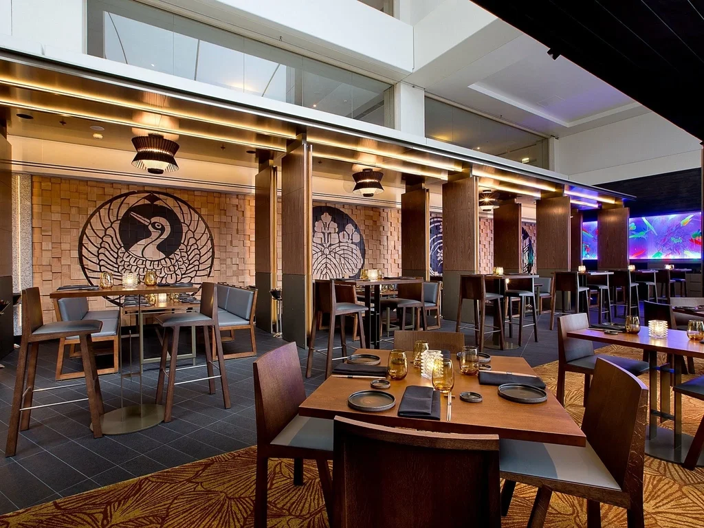 Elegant restaurant interior with wooden tables set with black plates, glasses, and candles, featuring Asian-inspired wall art and modern lighting.