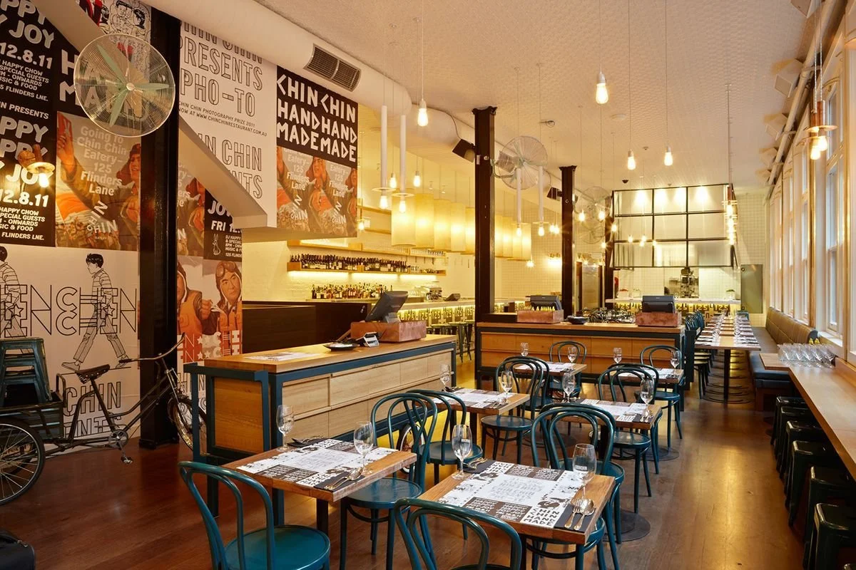 Interior of a modern restaurant with wooden tables and blue chairs, large windows on the right, wall decor with posters, a bicycle near the wall, and a bar area in the back with shelves of bottles and hanging light bulbs.