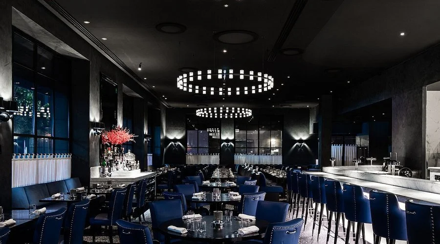 Interior of a modern upscale restaurant with dark walls, blue chairs, round tables set with glasses and utensils, and stylish lighting fixtures.