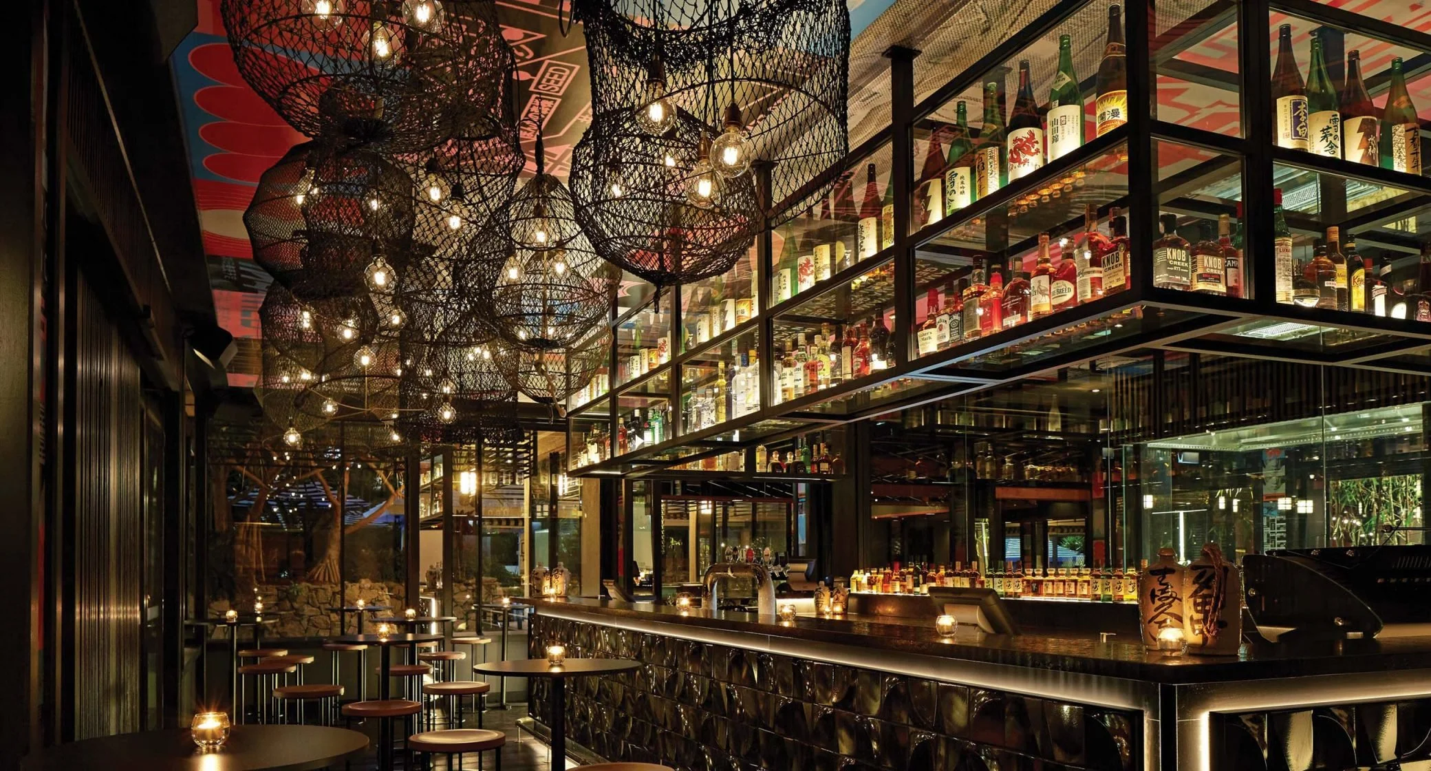 Interior of a modern bar or restaurant with hanging industrial-style wire basket light fixtures and a wall of liquor bottles behind the bar, illuminated warmly.
