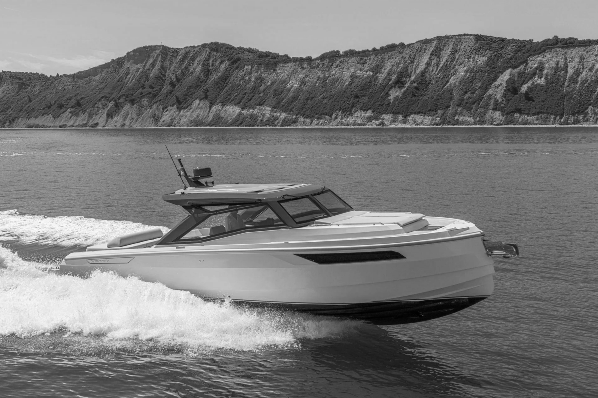 A white motor yacht cruising on a water body, with a steep hillside or cliff in the background.