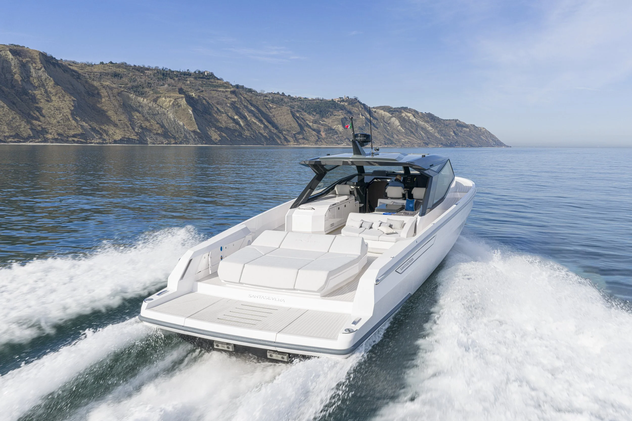 A white motor yacht speeding through open water with a backdrop of steep, rugged cliffs under a clear blue sky.