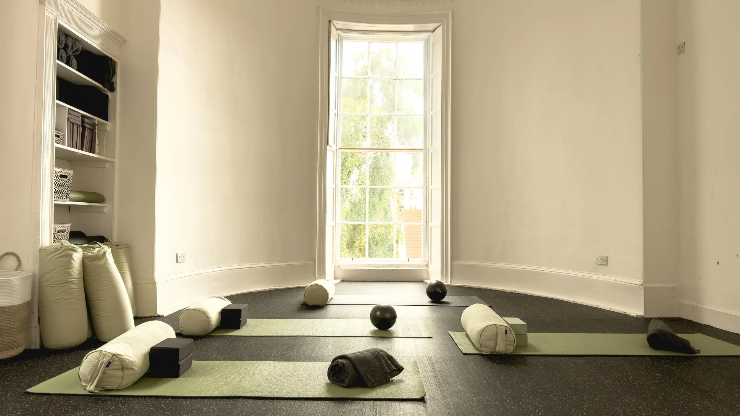 A yoga or meditation room with mats, bolsters, and exercise balls near a large window with open blinds, sunlight streaming in, and minimal decor.