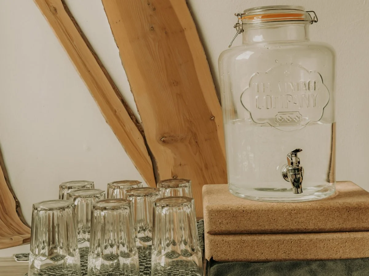 A clear glass water dispenser with a spigot, placed on a stack of two cork boards, next to several upside-down glass cups, against a wall with wooden paneling.