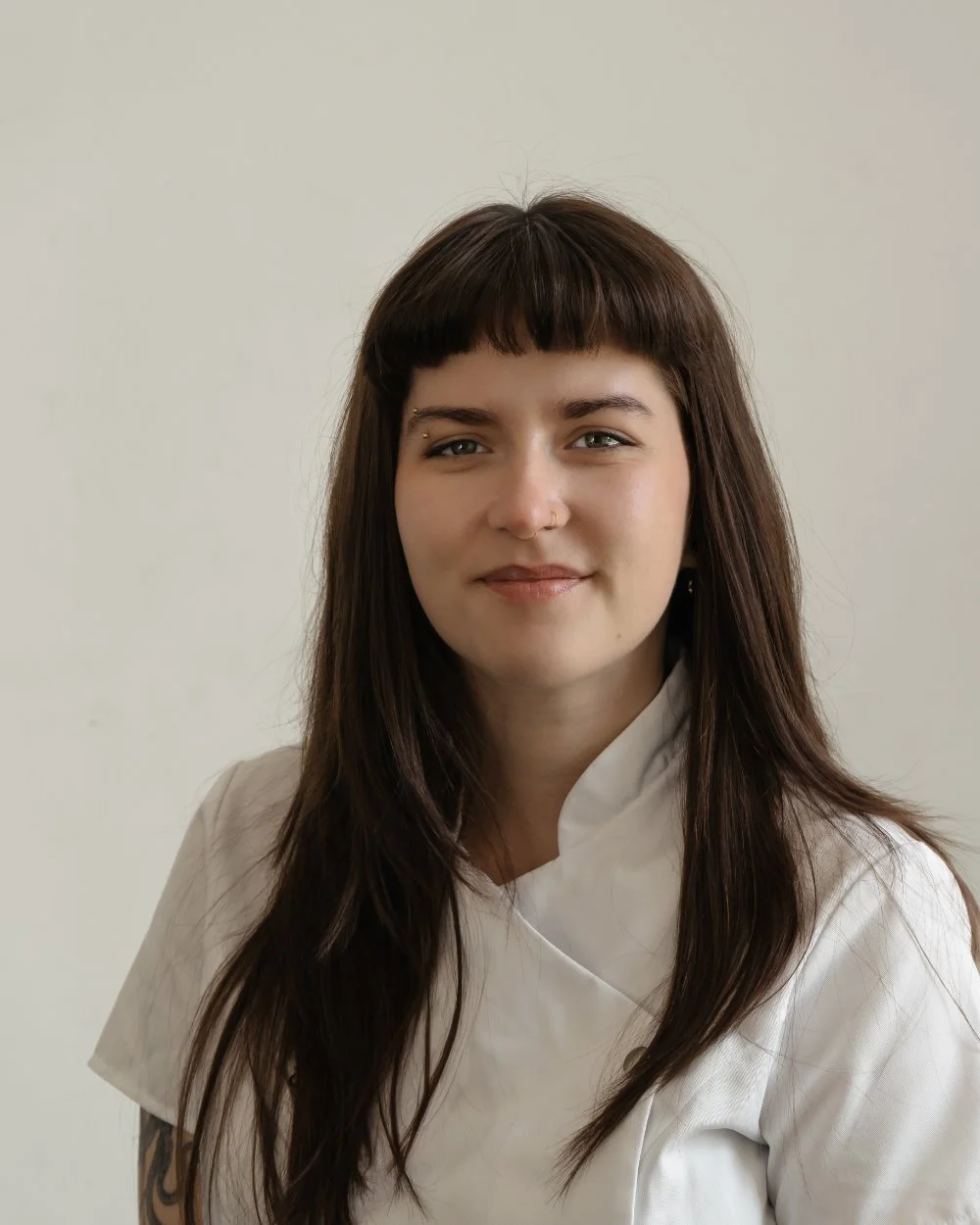 A young woman with long dark hair and bangs, wearing a white chef's coat, smiling softly against a plain light background.