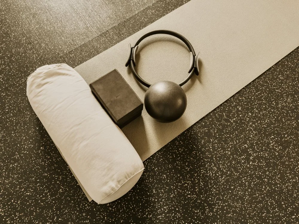 Fitness equipment including a yoga mat, black box, foam roller, medicine ball, and headphones on the floor.