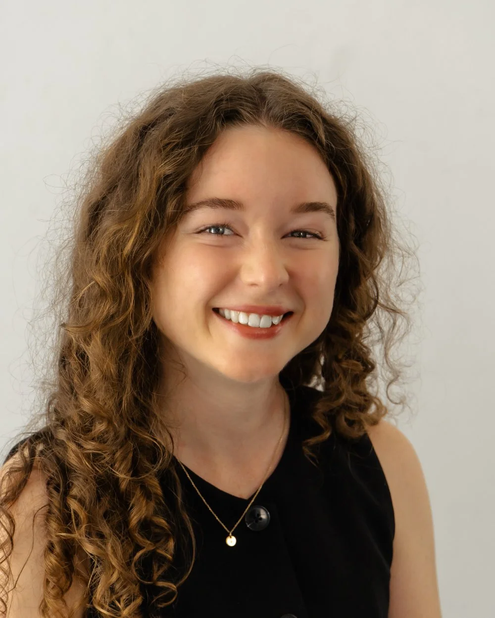 Close-up of a smiling woman with curly brown hair wearing a black sleeveless top and gold jewelry against a plain light background.