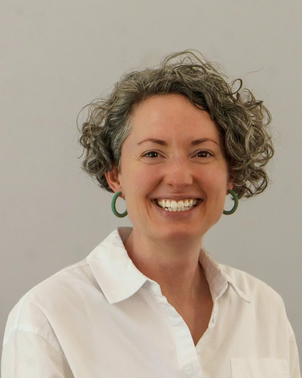 A woman with short curly gray hair, wearing a white shirt and green hoop earrings, smiling at the camera, against a plain light background.
