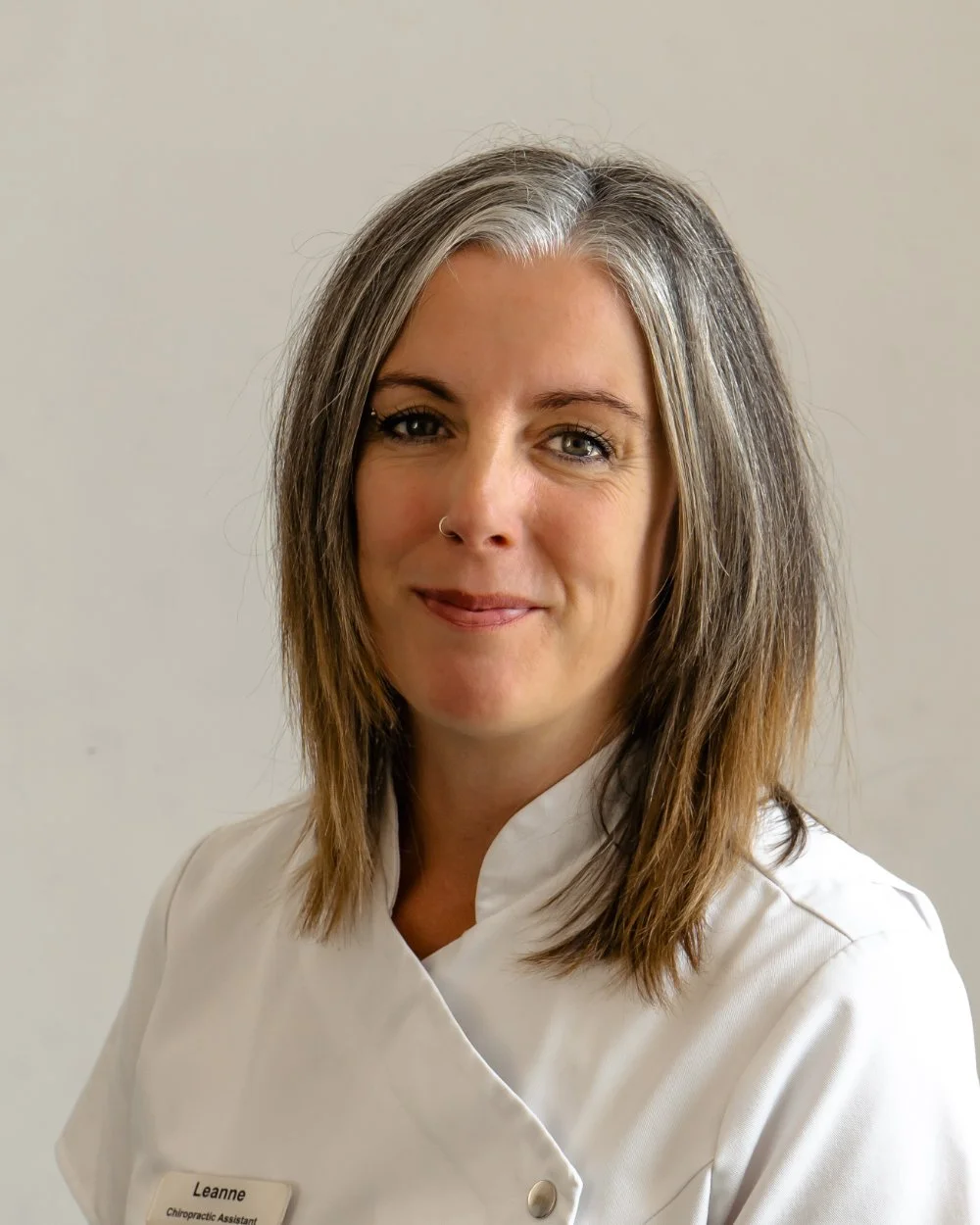 A woman with shoulder-length gray and brown hair, wearing a white medical uniform with a name tag that reads "Leanne" and "Chiropractic Assistant." She is smiling slightly against a plain light-colored background.