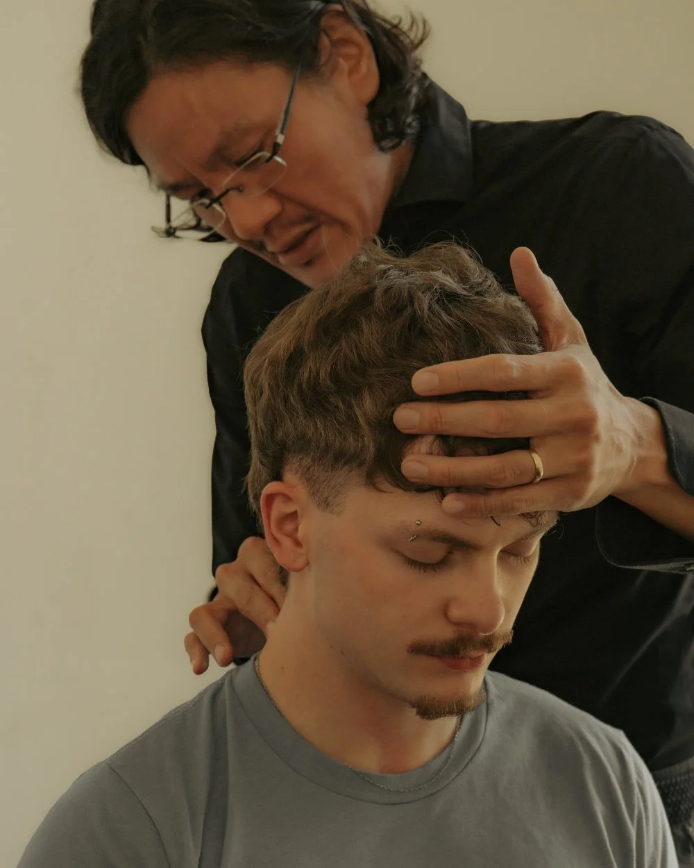 A man with glasses is holding a young man's head gently, his eyes closed, during a moment of prayer or meditation, in a room with a plain light-colored wall.