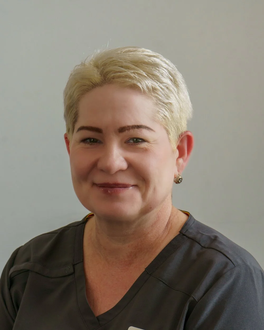 Close-up of a smiling woman with short blond hair, wearing earrings and a dark gray uniform.