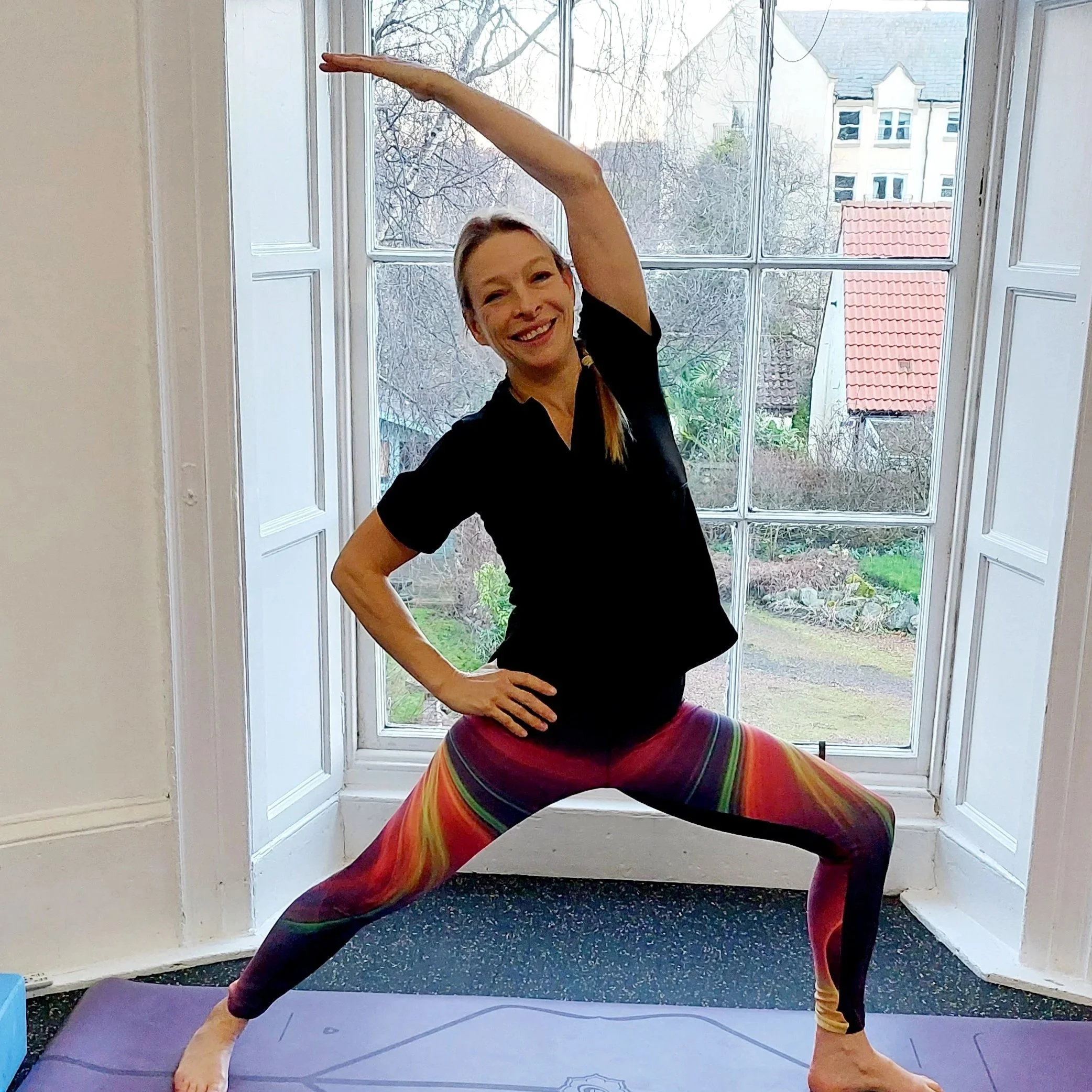 A woman in colorful striped leggings and a black top performs a yoga pose in front of a large window with a view of trees and neighboring buildings, standing on a purple yoga mat.