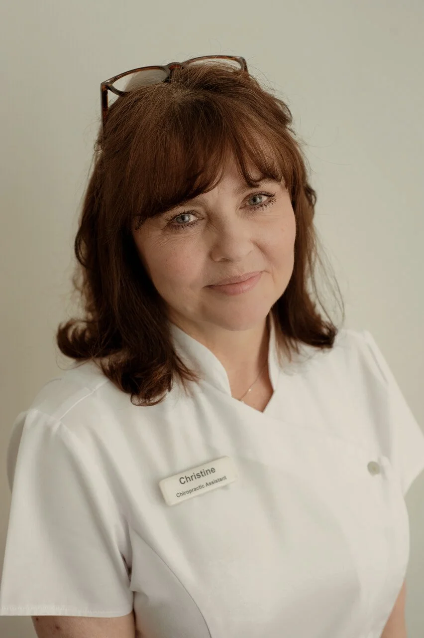 Portrait of a woman wearing a white medical uniform with a name tag that reads "Christine, Chiropractic Assistant," with glasses resting on her head, smiling softly.