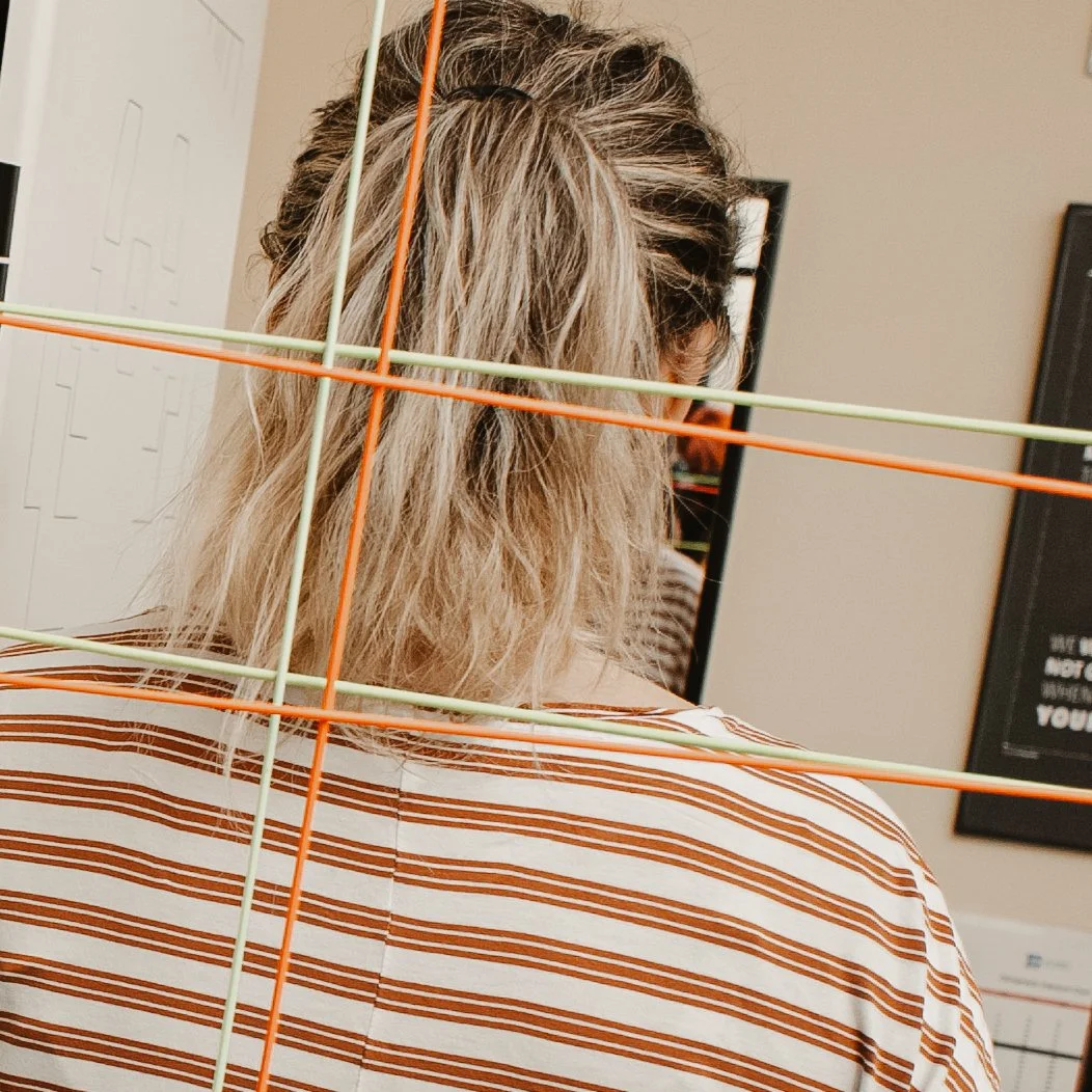 Person with wavy blonde hair wearing a striped shirt viewed from behind, with colorful plastic sticks arranged in a grid pattern in front of them, partially obscuring their head and shoulders, in an indoor setting with framed artwork on the walls.