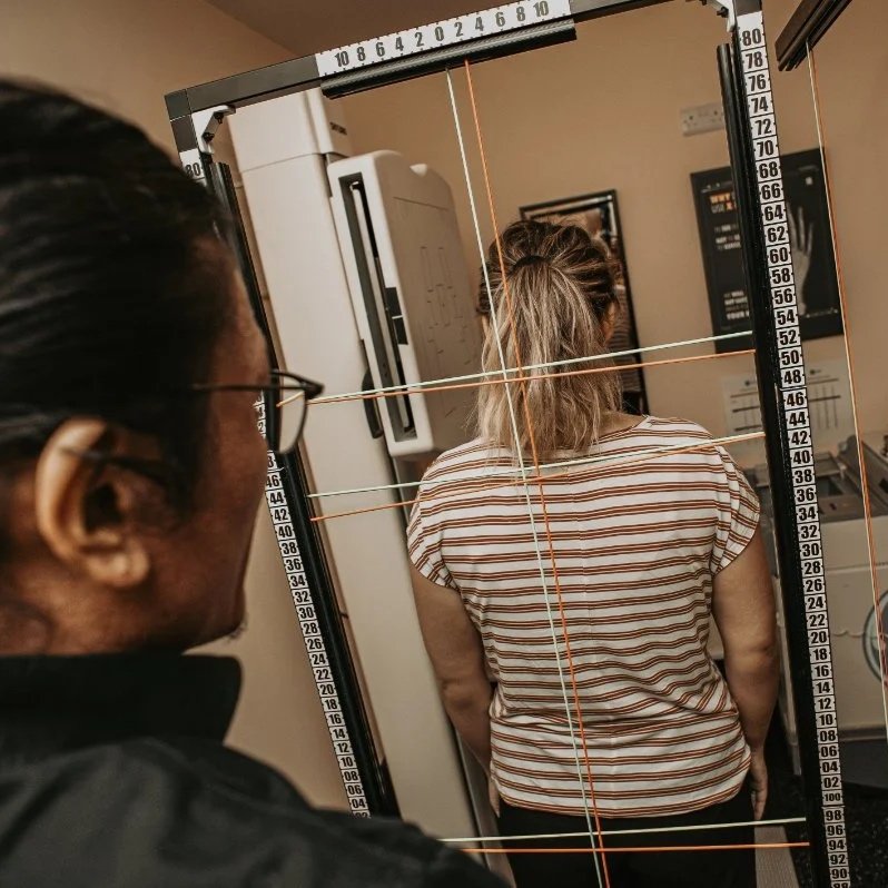 Woman with hair in a ponytail standing in front of a mirror, with a person observing her reflection. The mirror is part of a medical or scientific measurement setup with numbered scales and horizontal and vertical lines for alignment.