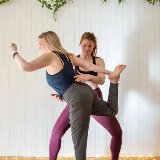 Two women practicing partner yoga in a studio with white panel walls and green plants.