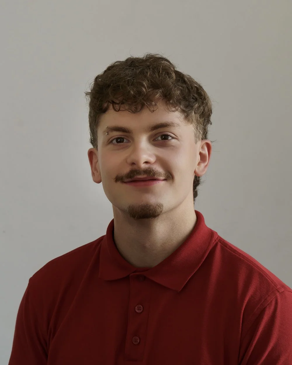 A young man with curly brown hair, a mustache, and a goatee wearing a red polo shirt, smiling softly, photo taken against a plain neutral background.