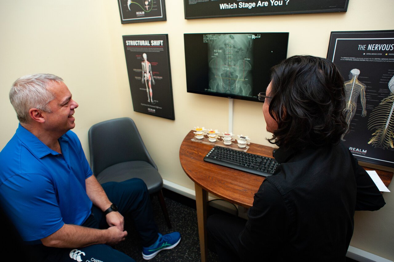 Dr Jon and a patient sitting and talking in an exam room with medical posters and X-ray images on the wall, a computer keyboard on the desk.