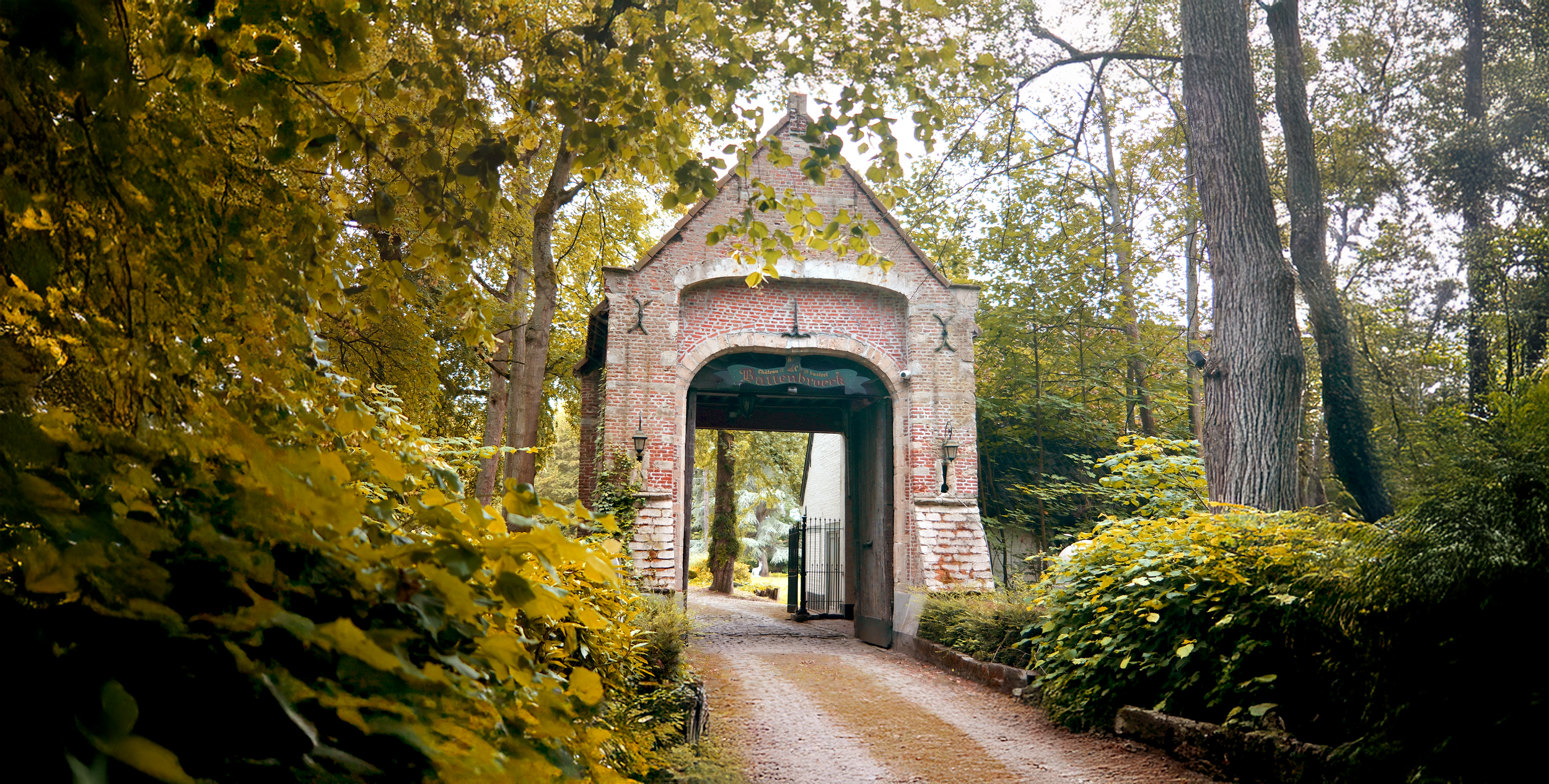 Een oude stenen poort met een boog, omringd door een bos met herfstkleurige bladeren. De poort leidt naar een pad in het bos.  | Kasteeldomein Battenbroek | Evenementenlocatie in Mechelen