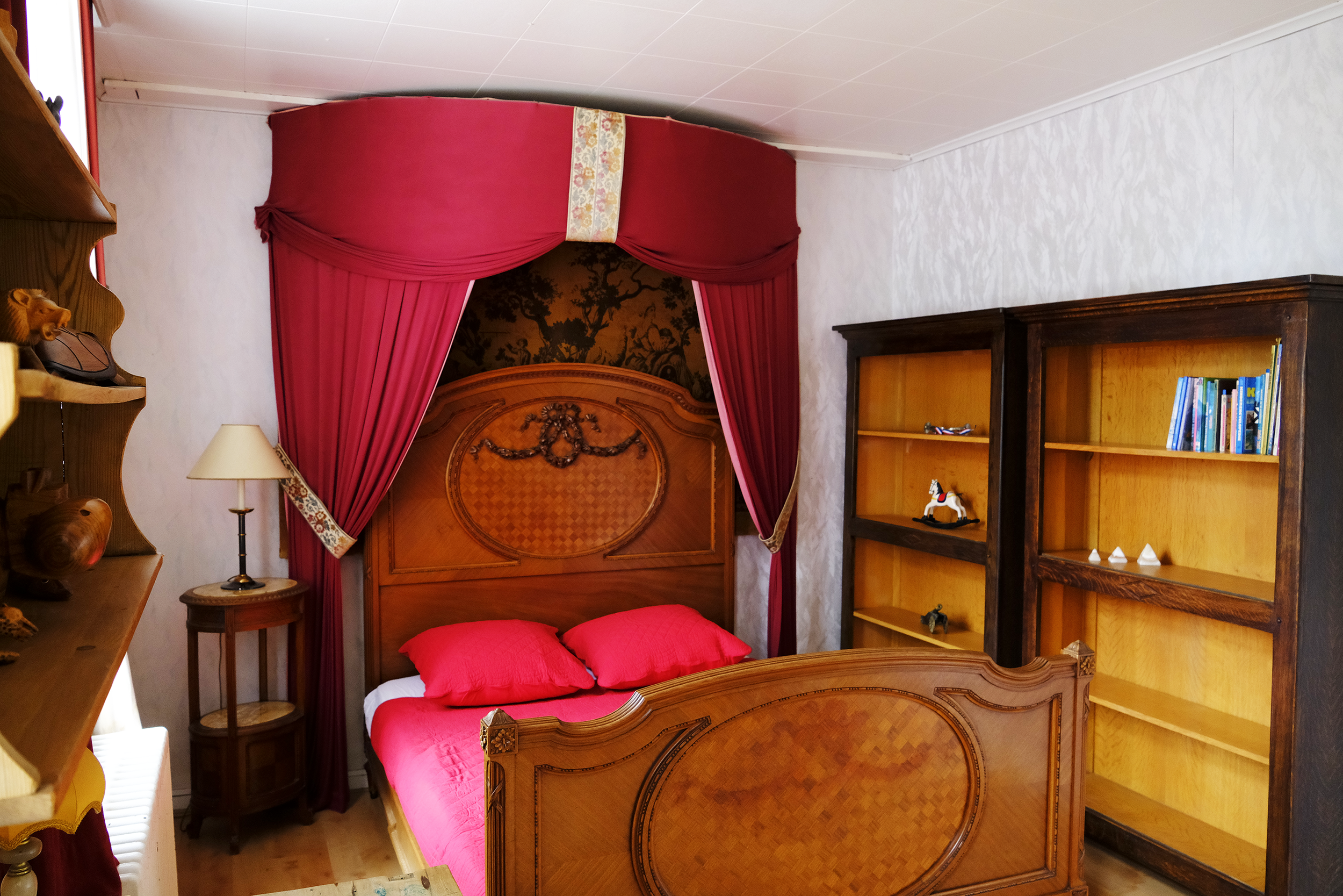 Een vintage houten bed met een bloemmotief en een geslage klamboe van rood linnen. Aan de zijde een nachtkastje met een kleine lamp en een tienerkamer met boekenplank en decoraties.  | Kasteeldomein Battenbroek | Evenementenlocatie in Mechelen