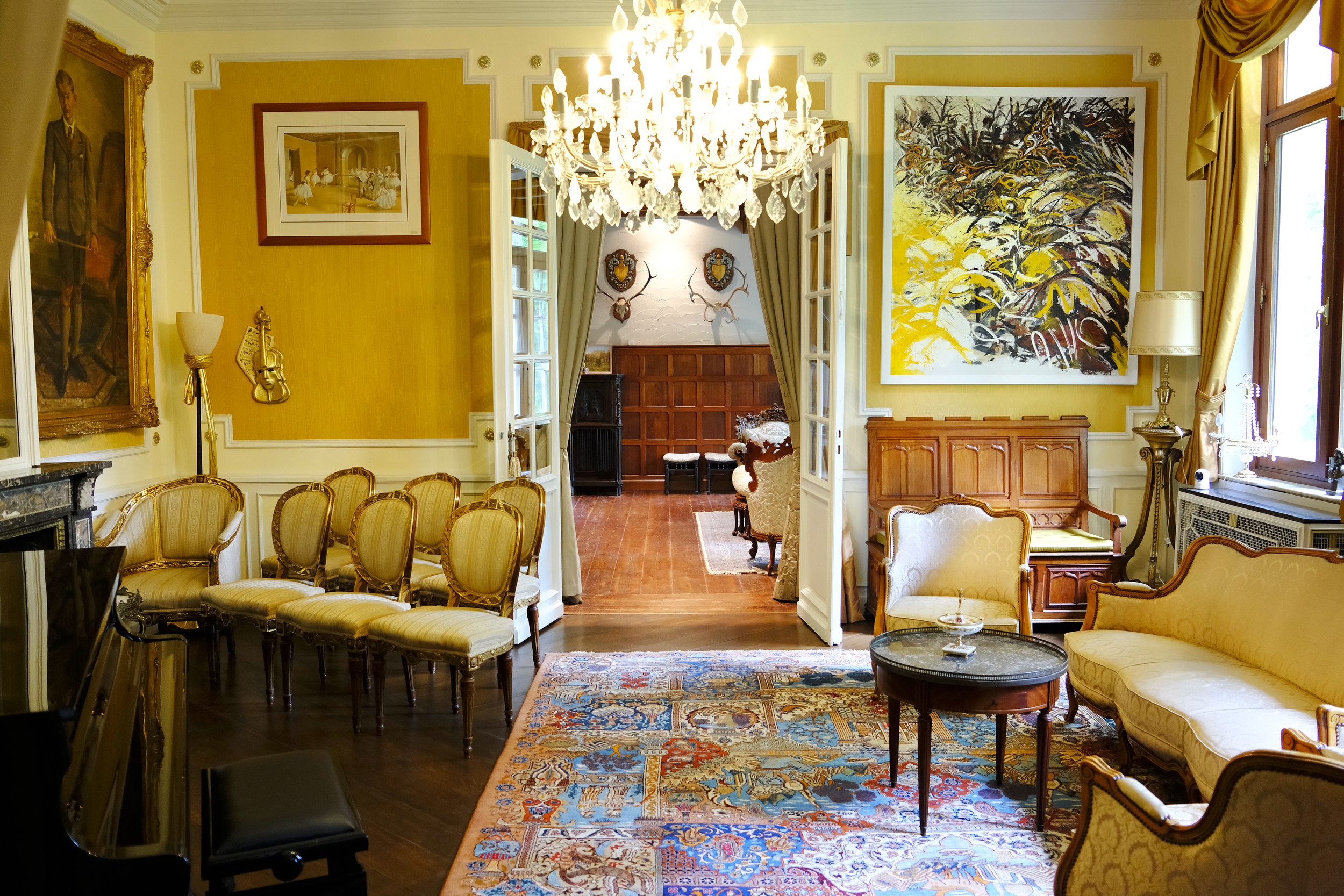 Interieur van een elegante, klassieke woonkamer met gele muren en diverse kunstwerken, waaronder een grote abstracte schildering en een portret, met een chandelier, oude meubels en een kleurrijk tapijt. | Kasteeldomein Battenbroek
