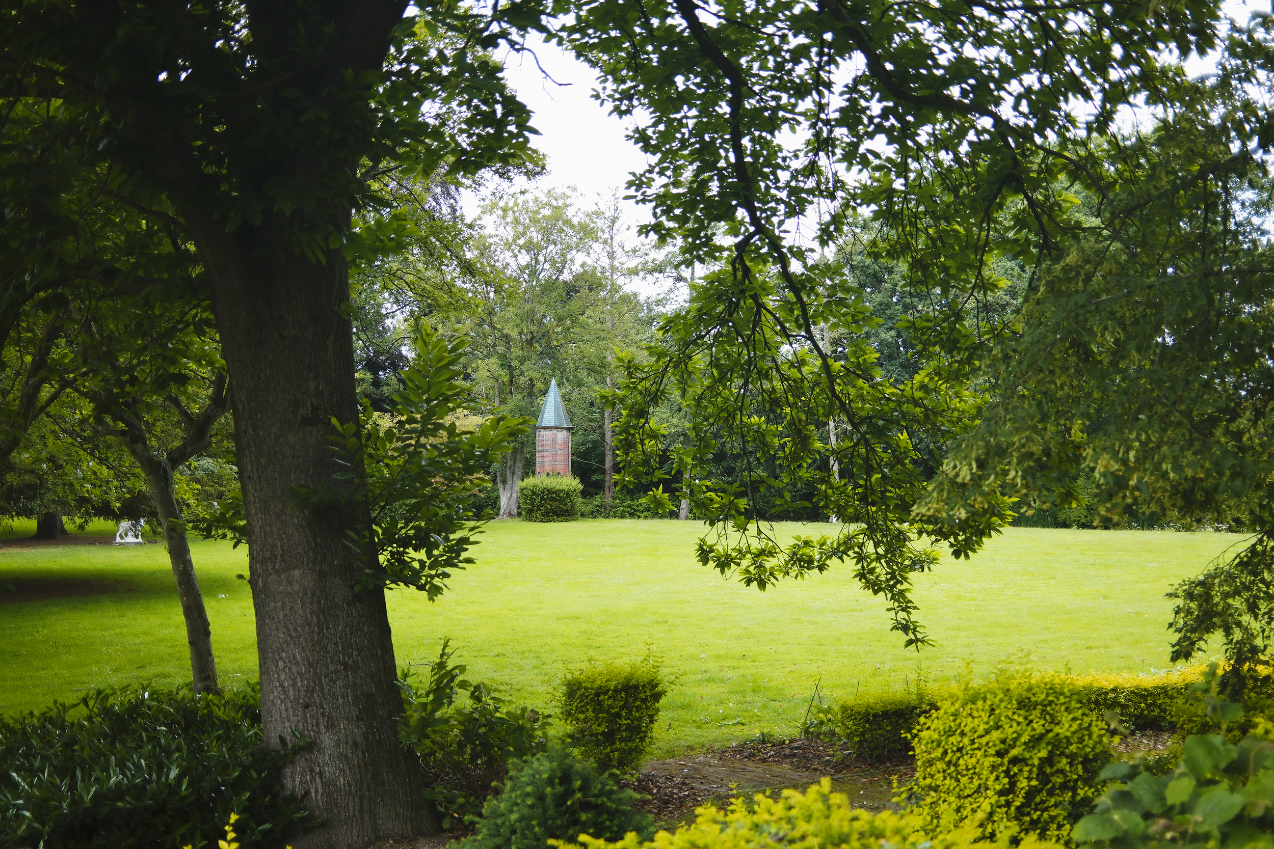 Een groen park met grote bomen, grasvelden en een toren met een puntig dak op de achtergrond.