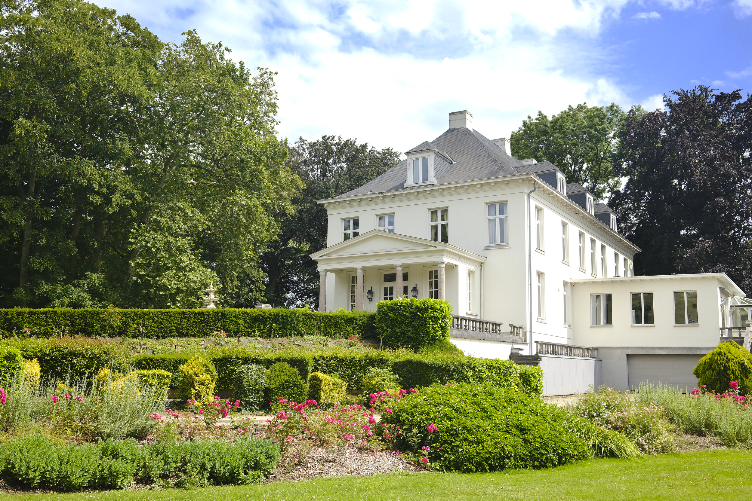 Een wit landhuis met meerdere verdiepingen, omlijst door groene bomen, een voor- en zijtuin met bloemen en een heg, onder een deels bewolkte blauwe lucht.