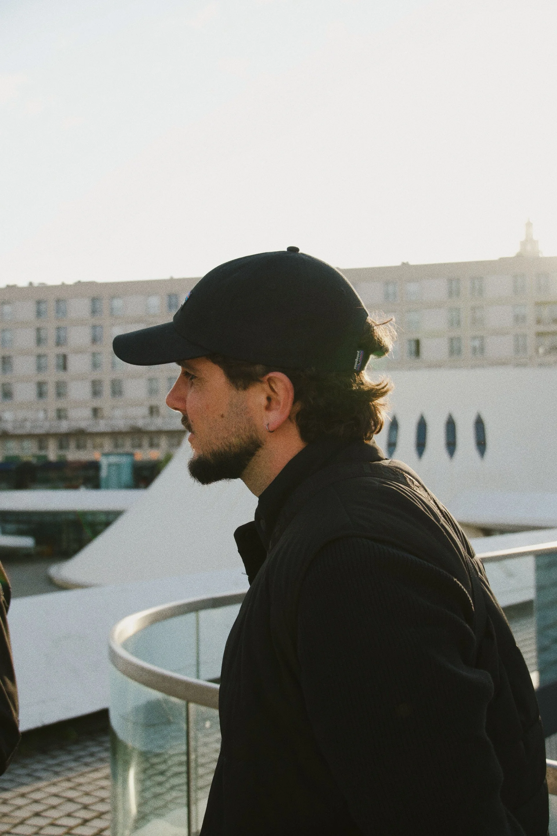Un homme portant une casquette noire et un manteau noir, vu de profil, se tenant près d'une barrière en métal sur un bateau ou une structure sur l'eau, avec un bâtiment résidentiel en arrière-plan lors du coucher du soleil.