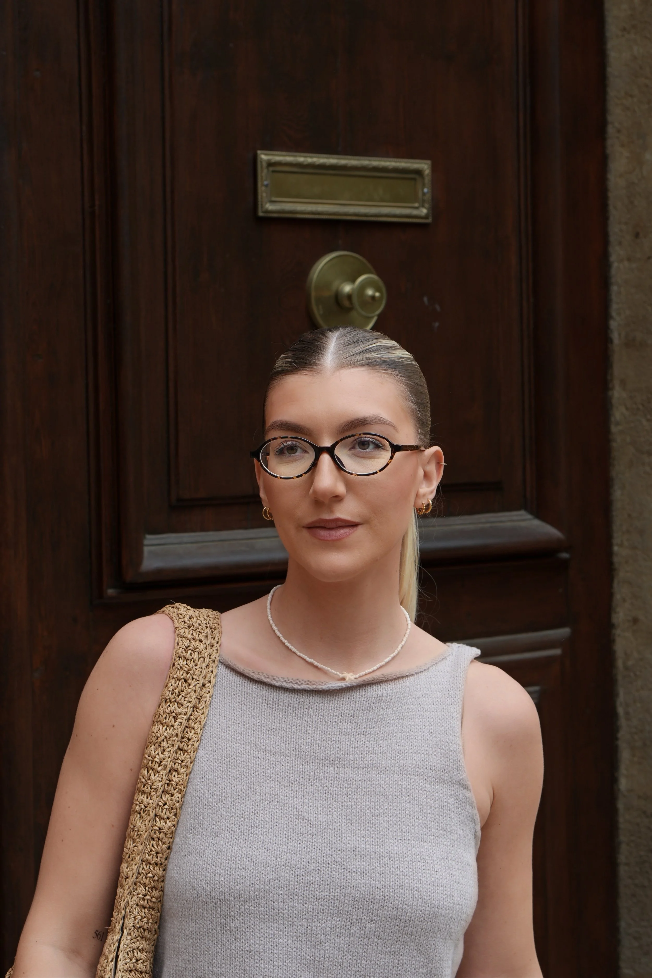 Jeune femme portant des lunettes, un bracelet en perles, un haut gris et un sac en fibres naturelles, posée devant une porte en bois avec une boîte aux lettres en laiton.