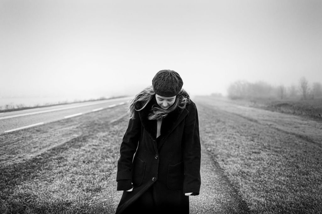 A woman wearing a coat and knit cap standing in a desolate outdoor area near the roadside, looking down with a somber expression.