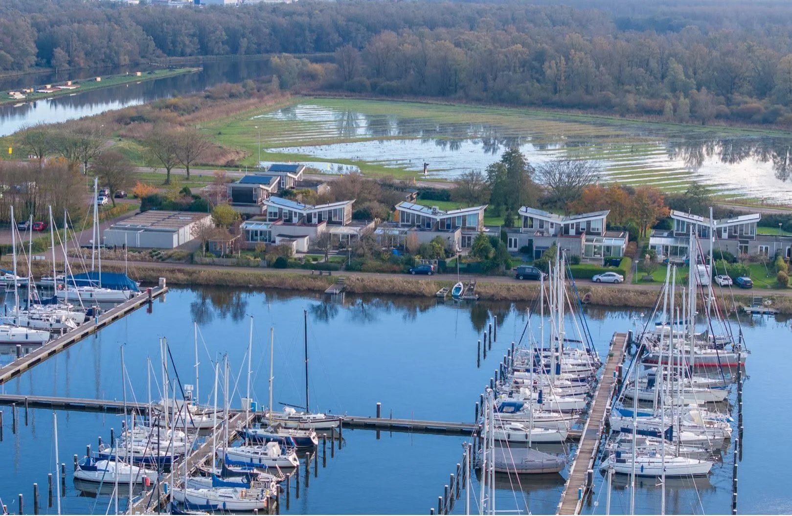 Marina with numerous sailboats docked at piers, modern houses in the background, and a river with wetlands and trees beyond.
