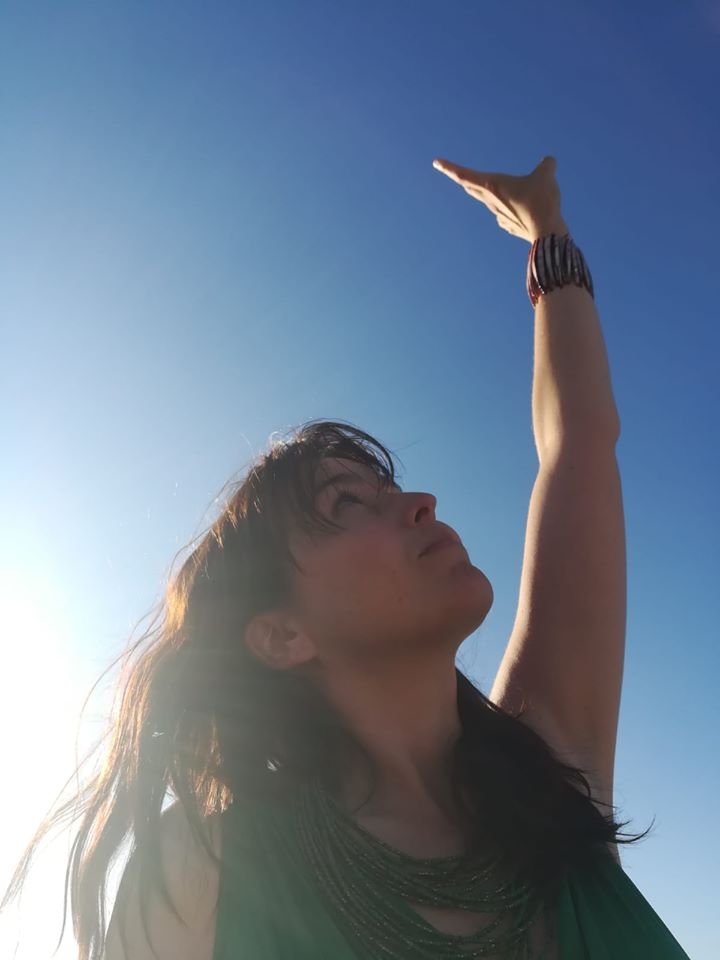 Woman standing outdoors against blue sky, raising her arm and reaching upward, with sunlight illuminating her profile.
