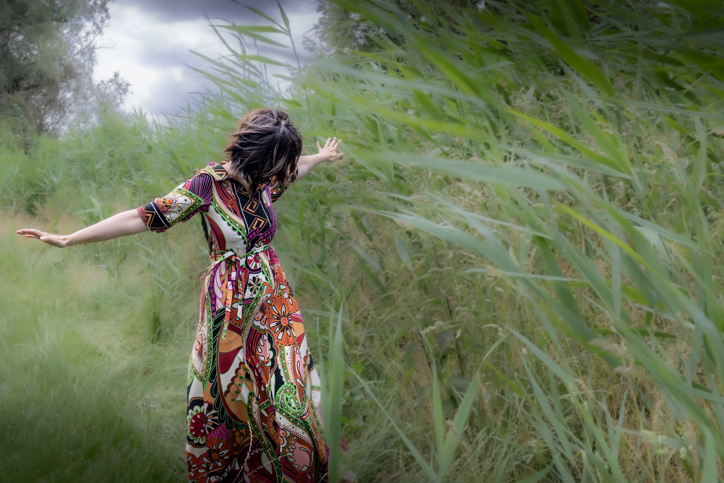 A woman wearing a colorful, patterned dress with outstretched arms in a green, outdoor natural setting with tall grass and trees.