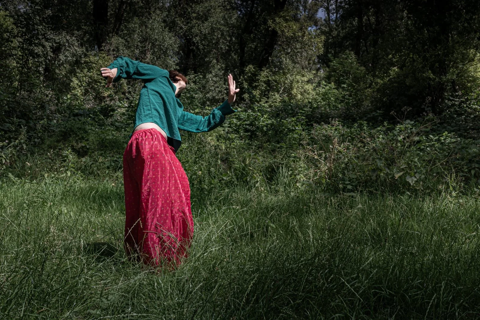 Person in a teal jacket and red skirt standing on grass in a forest, with arms raised and head turned away.