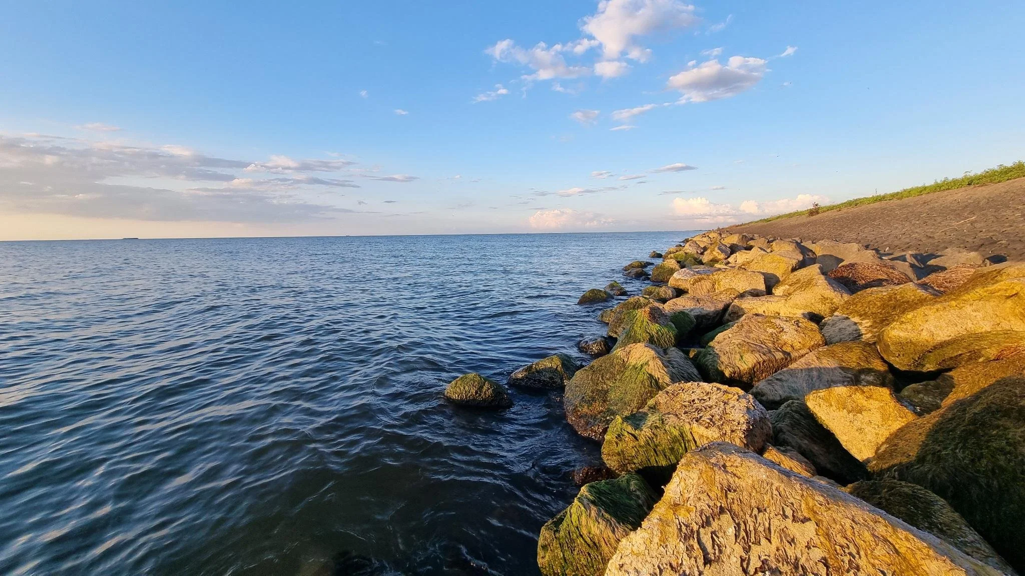 A view of a calm body of water with gentle ripples, a rocky shoreline with moss-covered stones on the right, and a clear sky with scattered clouds in the background during sunset or sunrise.
