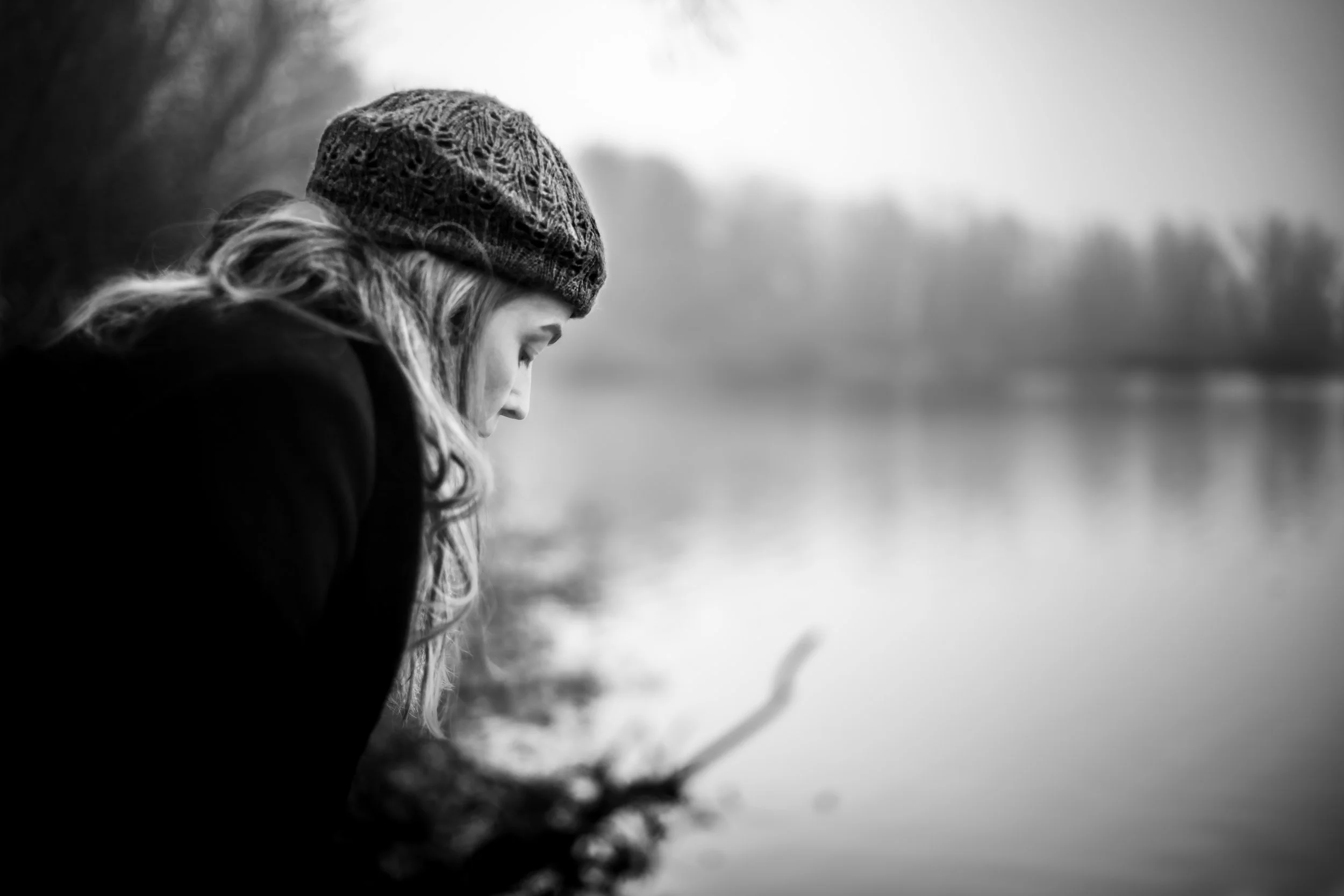 A black and white photo of a person with long hair wearing a knitted hat, looking down near a calm body of water surrounded by trees.