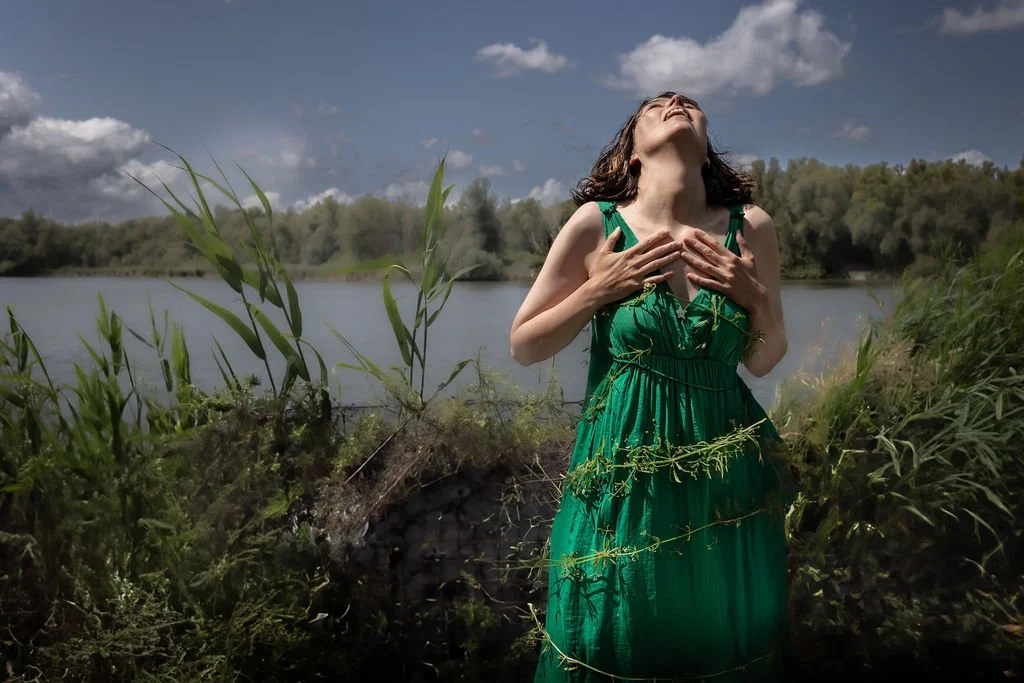 Woman in a green dress standing outdoors near a body of water, with her hands on her chest, head tilted back, and eyes closed, enjoying the outdoors on a sunny day.