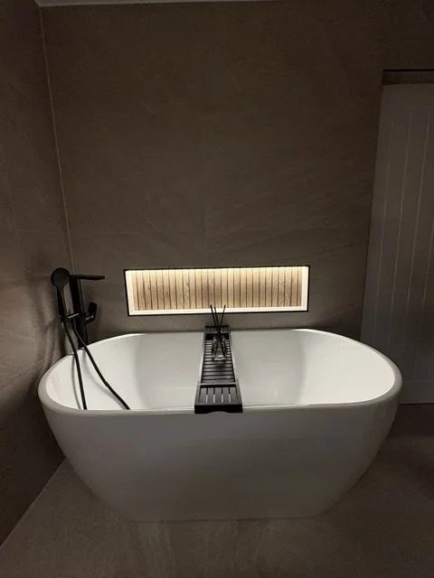 A modern white freestanding bathtub with a black bath caddy, located in a bathroom with beige tiled walls and a narrow, horizontal window behind it.