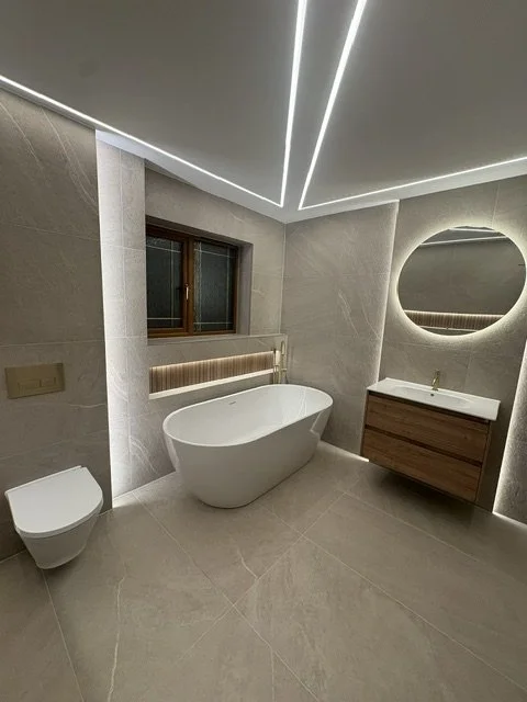 Modern bathroom with a white freestanding bathtub, a wall-mounted toilet, a wooden vanity with a sink, a round mirror, a small window, and sleek ceiling lighting.