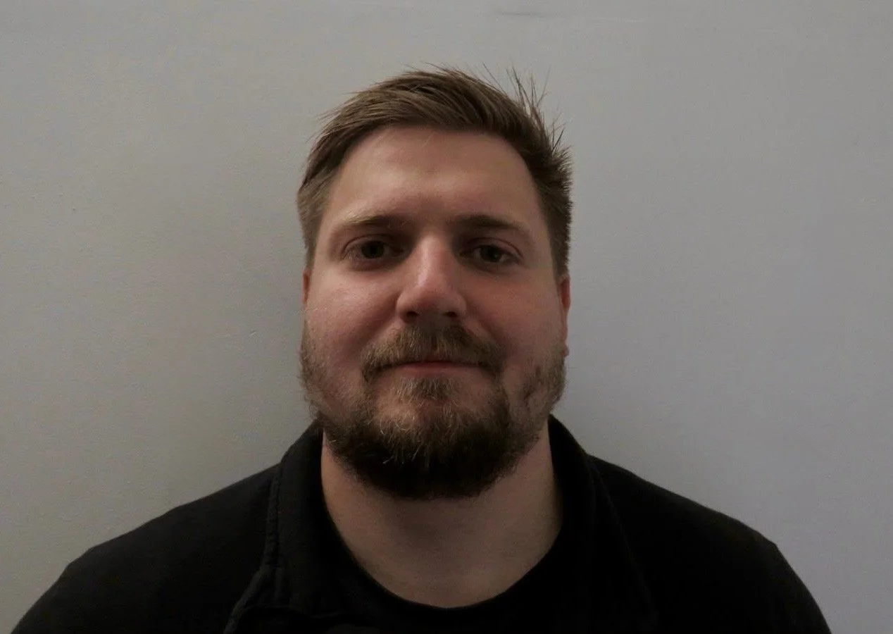 Portrait of a man with brown hair and beard, wearing a black shirt, looking at the camera against a light gray wall.